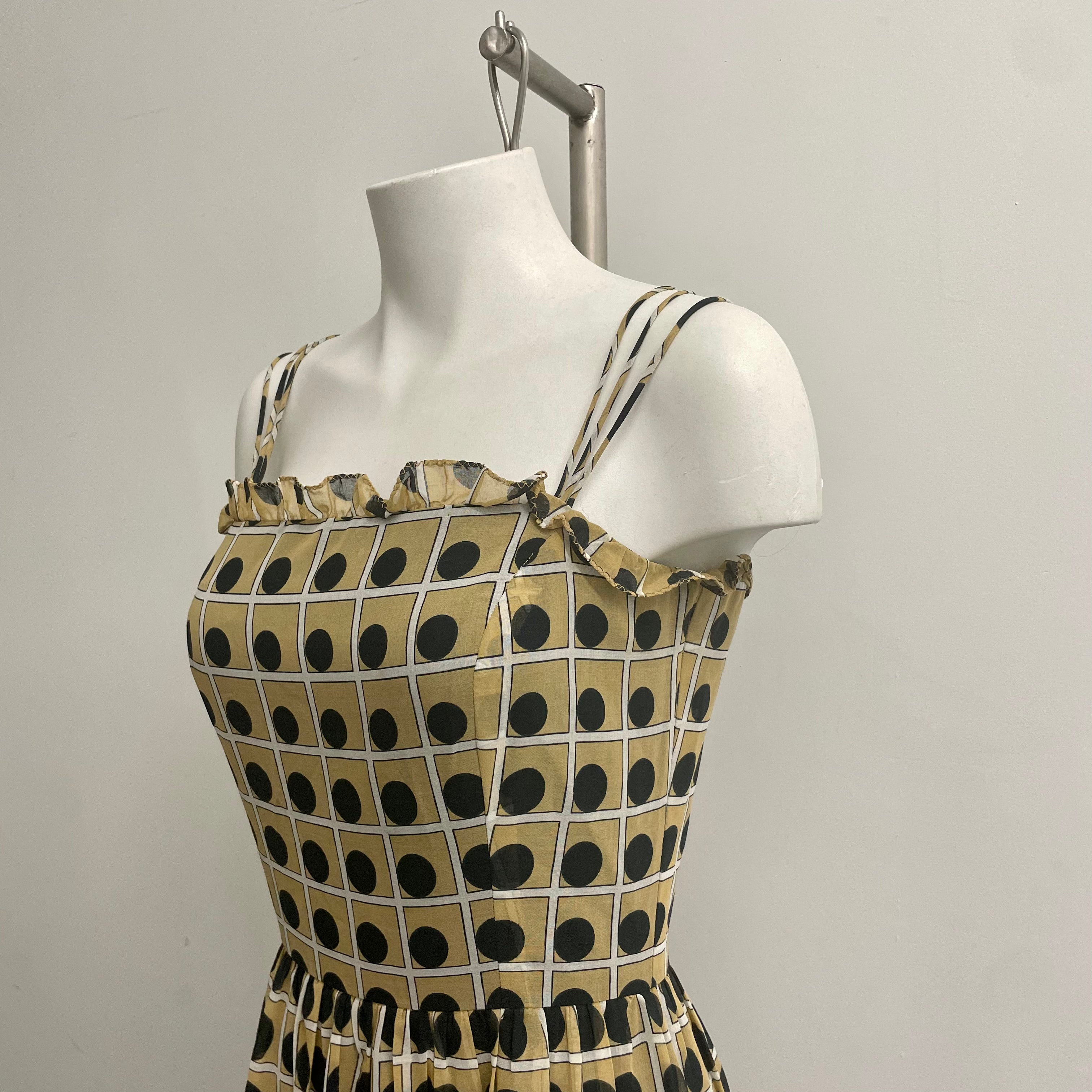 A sleeveless beige ocre (#e0cb92) dress with dark navy polka dots and a white grid pattern, with ruffled edge and 3 spaghetti straps displayed on a white mannequin torso  